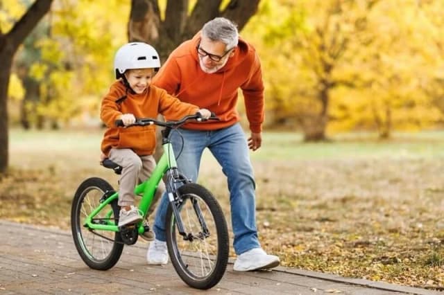 An older man walking alongside a young child, steadying the handlebars as the child rides a bicycle on a park path in autumn