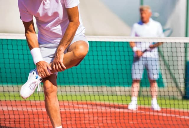 A tennis player on an outdoor court bending down to hold his lower leg and ankle after an injury